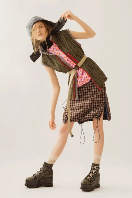 A woman wearing a gray bonnet, green vest, pink floral top, and plaid skirt strikes a pose in a fashion-forward shot in a studio setting.