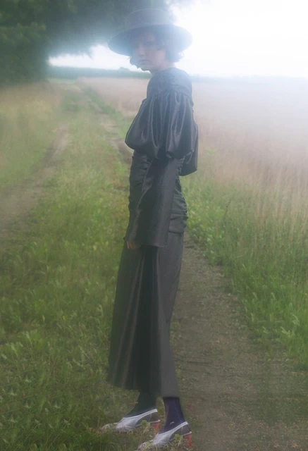 A female model stands on a dirt path in a rural area, wearing a dark dress with exaggerated sleeves, white heels, and a wide-brimmed hat.