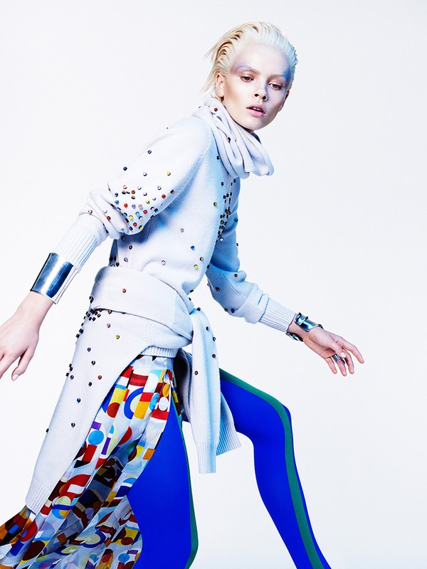 Fashion editorial shot of a blonde adult female wearing a colorful avant-garde outfit and metal cuff bracelets against a bright background.