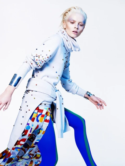 Fashion editorial shot of a blonde adult female wearing a colorful avant-garde outfit and metal cuff bracelets against a bright background.