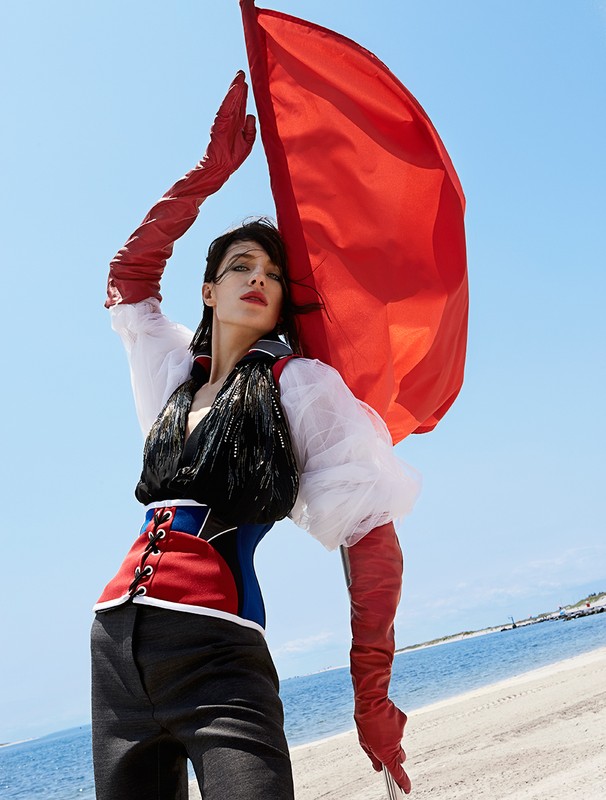 Fashion editorial shot: Woman on beach holding red flag, wearing corset, long gloves, black pants, white frilly top against blue sky.