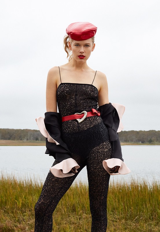 Young woman poses in a black lace bodysuit with a red beret, and a red heart-shaped belt standing outside with a cloudy sky backdrop.