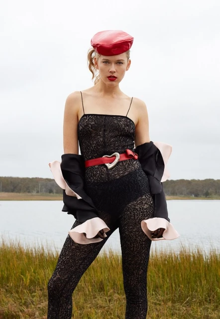 Young woman poses in a black lace bodysuit with a red beret, and a red heart-shaped belt standing outside with a cloudy sky backdrop.