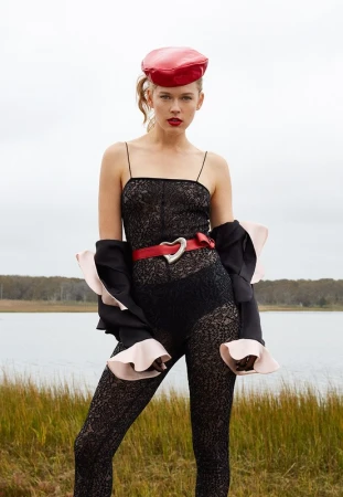 Young woman poses in a black lace bodysuit with a red beret, and a red heart-shaped belt standing outside with a cloudy sky backdrop.