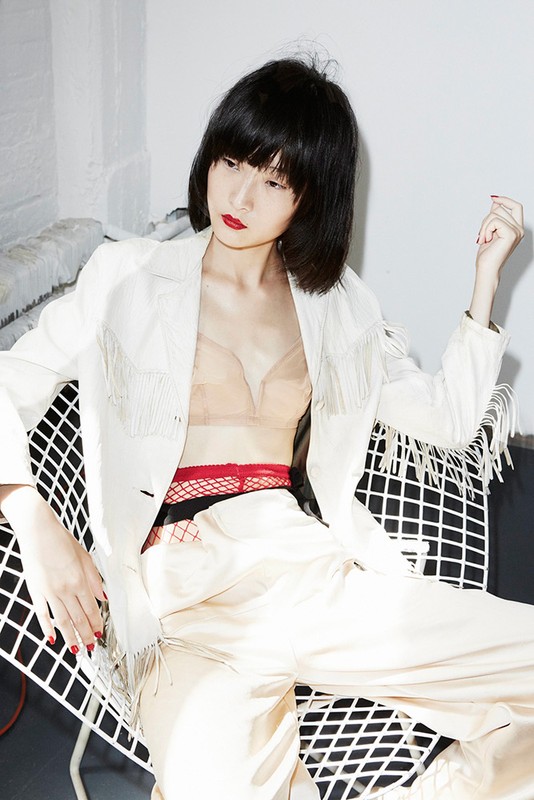 Elegant woman in a white fringe jacket and satin pants poses in a wire chair against a white wall. Fashion editorial shot.