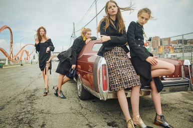 Four female models posing with a vintage red car in an outdoor street setting with a roller coaster in the background