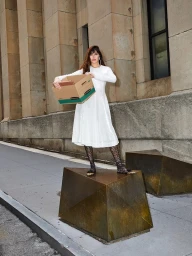 Woman in white midi dress holding cardboard box standing on brass block against stone building