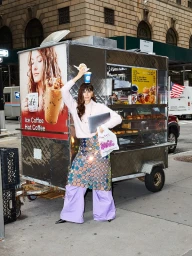 Woman in pink sweater and sequin skirt balancing coffee and pastry on her head in front of a food cart.