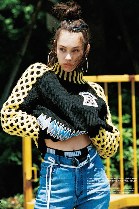 A young woman with a top-knot bun wearing a black and yellow knit sweater and blue jeans outdoors