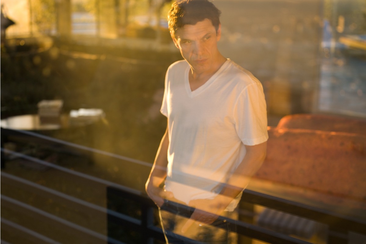 Adult man in a white t-shirt stands outdoors with hands in pockets and a serious expression, with blurred golden light in background.