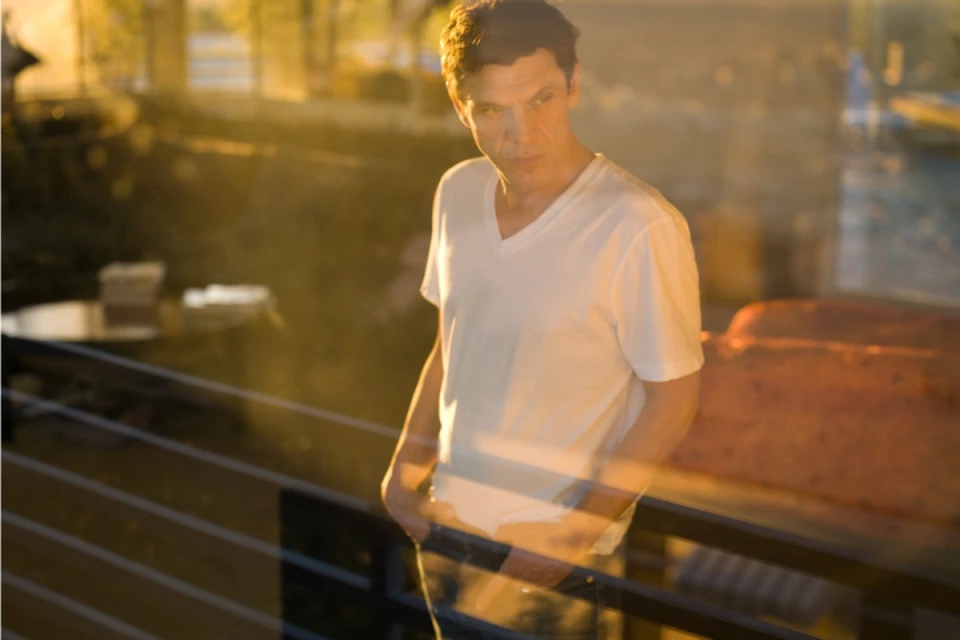 Adult man in a white t-shirt stands outdoors with hands in pockets and a serious expression, with blurred golden light in background.