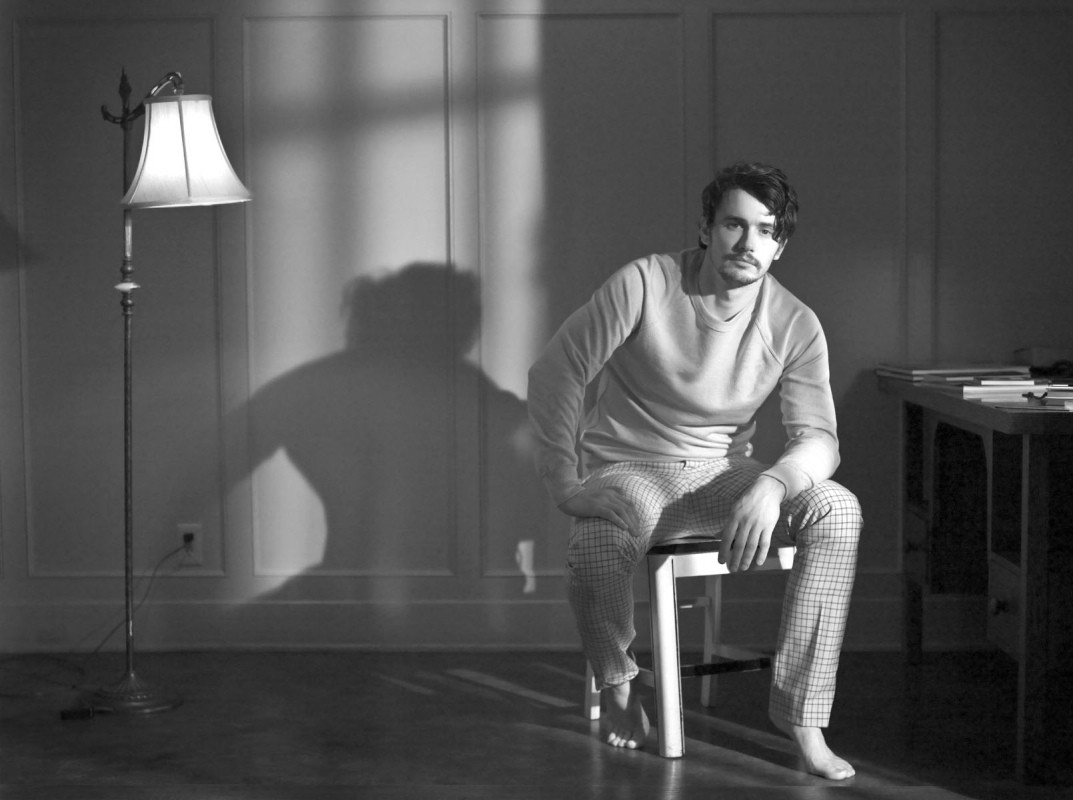 Monochrome portrait of James Franco seated on a stool indoors with a floor lamp and a dramatic shadow on the paneled wall behind him.