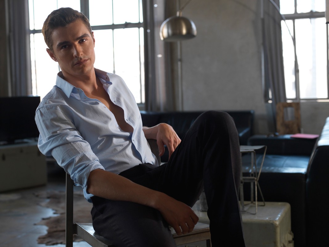 Portrait of a young adult male seated indoors, wearing an unbuttoned light blue shirt and dark pants, with natural light from a window.