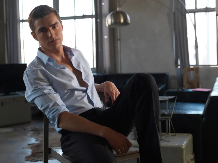 Portrait of a young adult male seated indoors, wearing an unbuttoned light blue shirt and dark pants, with natural light from a window.