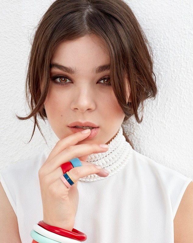 Close-up portrait of a woman with brown hair in a white top, wearing colourful rings and bangles, against a white background.