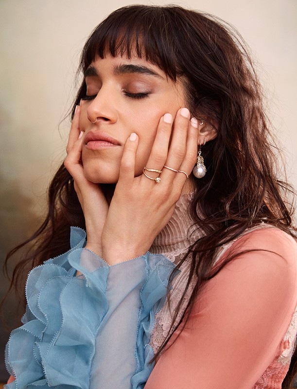 Portrait of a brunette woman with bangs and delicate gold jewelry, her hands gently touching her face in a soft, neutral-toned setting.