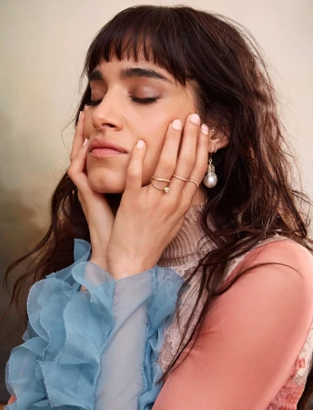 Portrait of a brunette woman with bangs and delicate gold jewelry, her hands gently touching her face in a soft, neutral-toned setting.