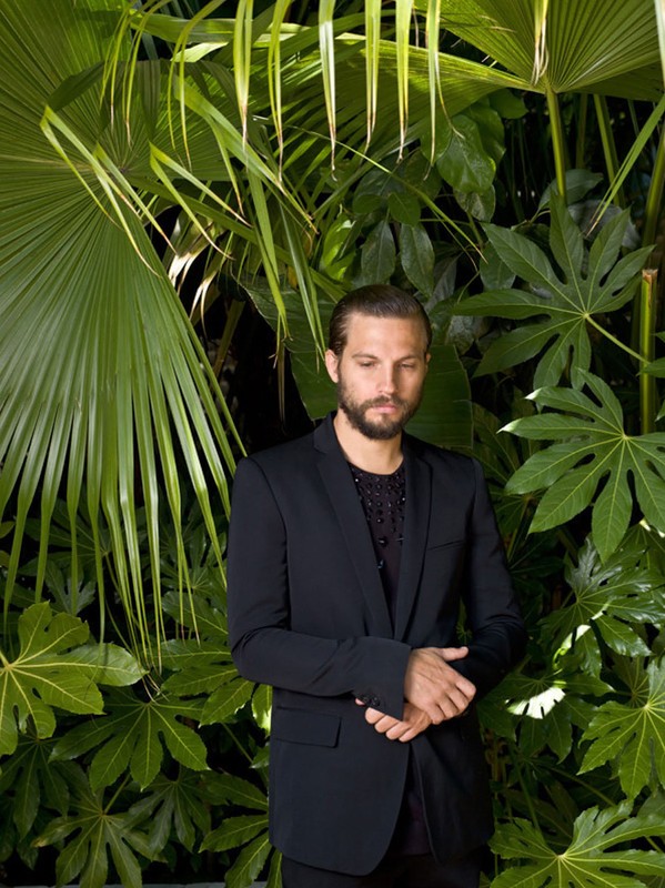 A man in a dark suit poses against a backdrop of lush green tropical foliage, looking downward in an outdoor setting with natural lighting.