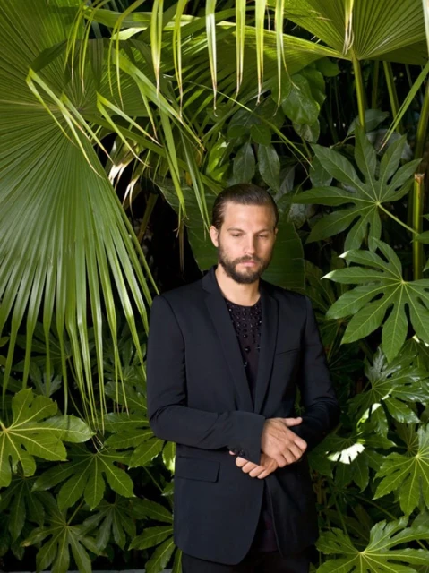 A man in a dark suit poses against a backdrop of lush green tropical foliage, looking downward in an outdoor setting with natural lighting.