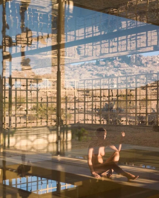 Reflection of a woman sitting indoors by a window, overlooking an arid outdoor landscape with desert vegetation and rocky hills.