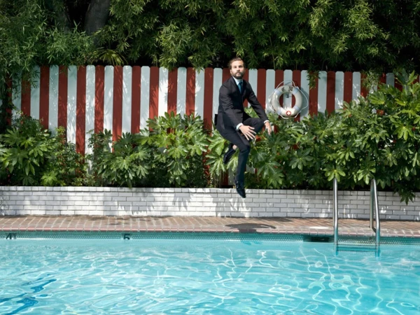 A man in a suit mid-air near a blue swimming pool, backed by red and white striped fence, green plants and a white life preserver.