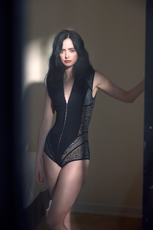 Brunette woman poses in a black bodysuit with lace panels, standing indoors next to a doorway with diffused light and shadows.