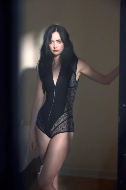 Brunette woman poses in a black bodysuit with lace panels, standing indoors next to a doorway with diffused light and shadows.
