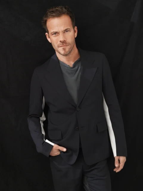 Portrait of a handsome man in a dark blazer and gray t-shirt, hand in pocket, against a dark background. Head and shoulders shot.