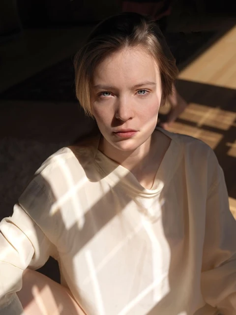 Indoor portrait of a young adult female with blonde hair and blue eyes, posed in direct sunlight with shadows, wearing a cream-colored top.