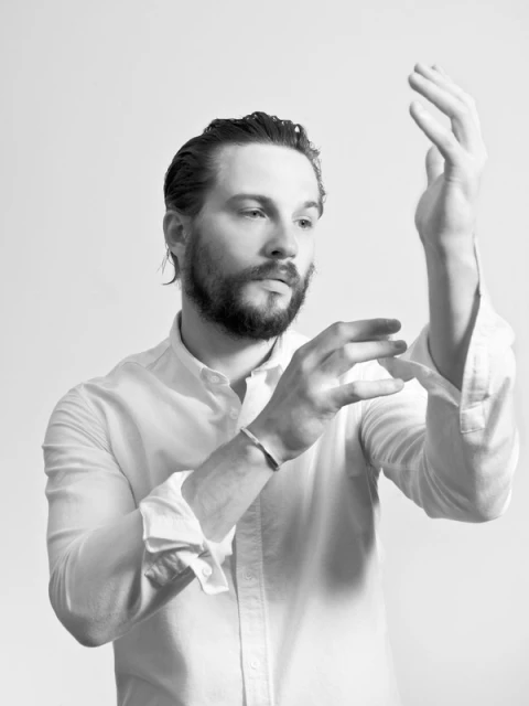 Portrait of an adult male with a beard wearing a white button-down shirt, adjusting his sleeves against a neutral background.