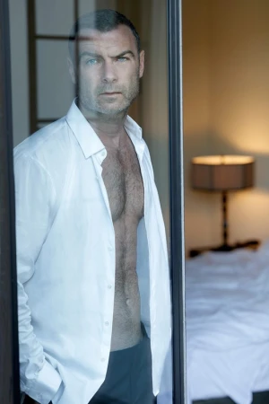 Portrait of actor Liev Schreiber standing in a doorway wearing an unbuttoned white shirt and dark trousers.