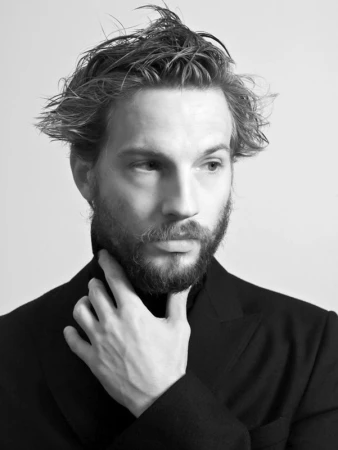 Monochrome portrait of a bearded man with tousled hair wearing a dark jacket, touching his chin.
