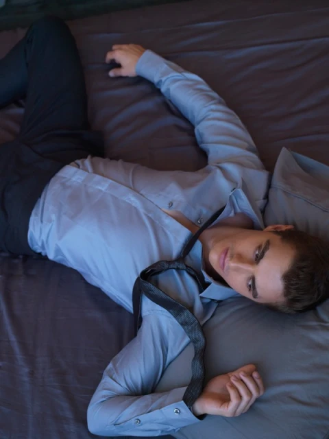 An adult male laying on a gray bed with an unbuttoned blue dress shirt and an untied tie with a gray pillow in the background.