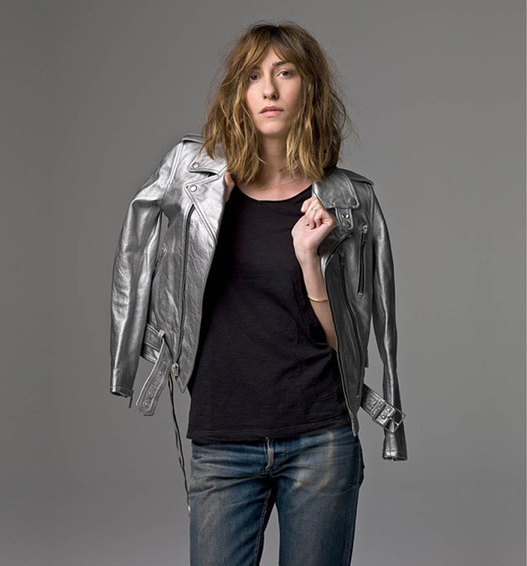 Woman in a black t-shirt and blue jeans poses in a photo studio wearing a silver metallic motorcycle jacket over her shoulder.