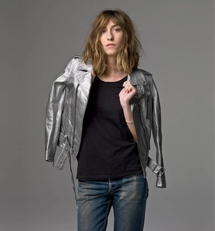 Woman in dark t-shirt and blue jeans wearing a metallic silver leather jacket against a plain grey background