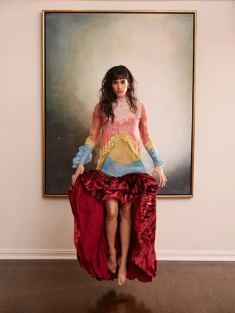 Woman in a colorful multi-layered dress hovering in mid-air in front of a large framed abstract painting.