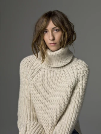 Woman with shoulder-length wavy brown hair wearing a chunky cream ribbed turtleneck sweater against a solid grey background