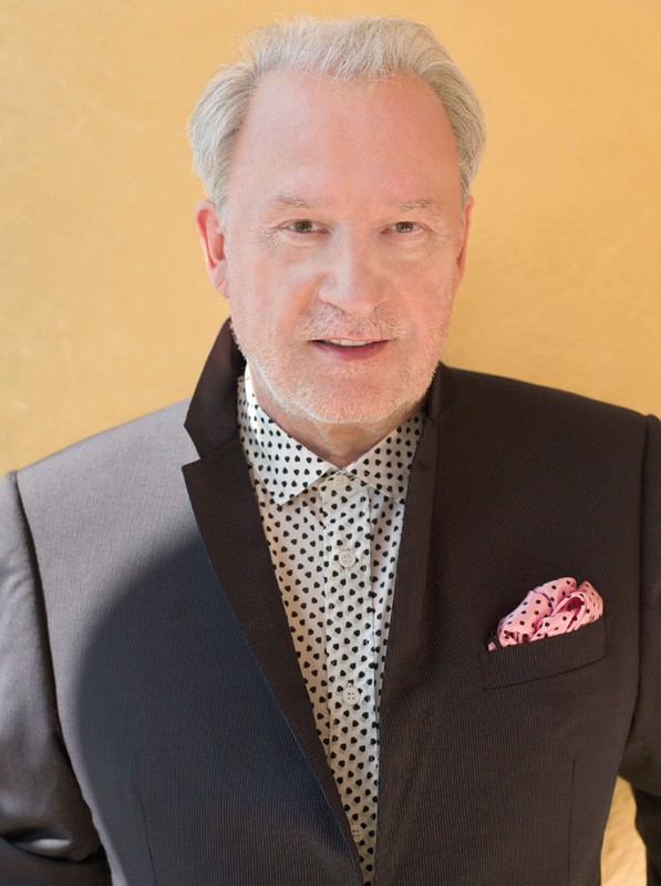 Portrait of a mature adult male in a suit with a patterned shirt, posing in front of a light yellow-tan background, studio portrait.