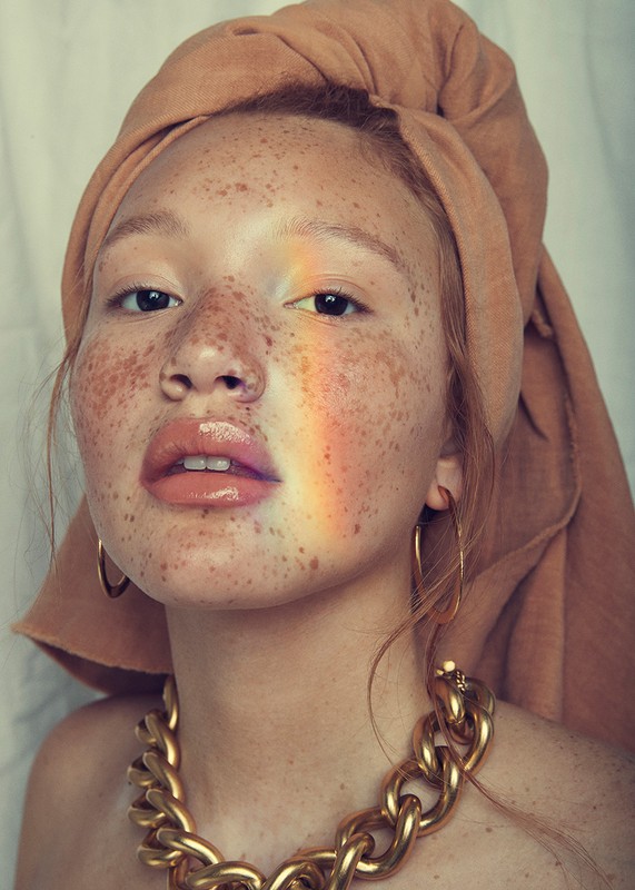Close-up portrait of a young adult woman with freckles, wearing a headscarf and gold necklace, featuring a rainbow light effect.