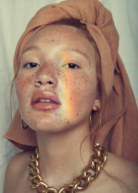 Close-up portrait of a young adult woman with freckles, wearing a headscarf and gold necklace, featuring a rainbow light effect.