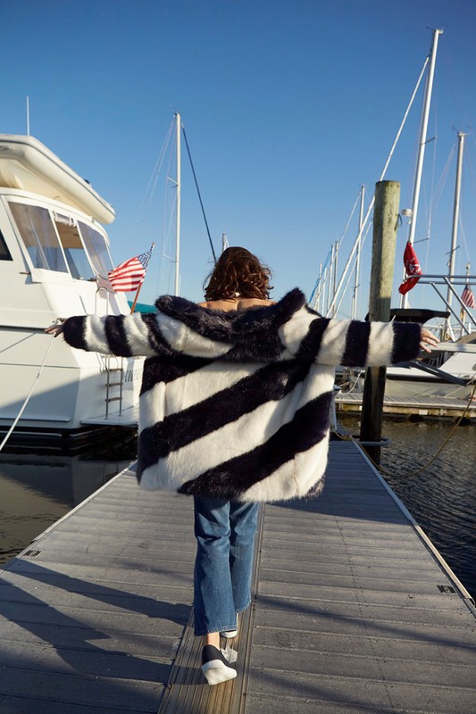 A woman with open arms walks on a pier toward boats under a blue sky, wearing a striped coat, blue jeans, and unique platform shoes.