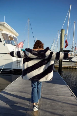 A woman with open arms walks on a pier toward boats under a blue sky, wearing a striped coat, blue jeans, and unique platform shoes.