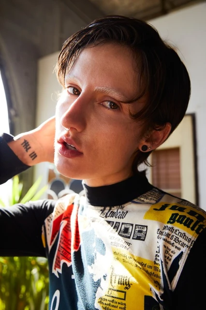 Close-up shot of a person with short brown hair wearing a patterned top, indoor setting with natural light coming from a window.
