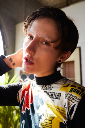 Close-up shot of a person with short brown hair wearing a patterned top, indoor setting with natural light coming from a window.