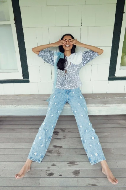 A young woman with long dark hair and blue jeans with white pom poms sits on a porch, covering her eyes with her hands, smiling.