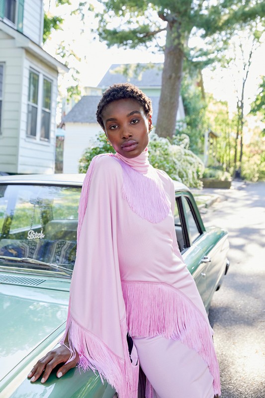 A stylish woman in a pink fringe dress poses against a vintage car in a suburban setting, bathed in soft natural light.