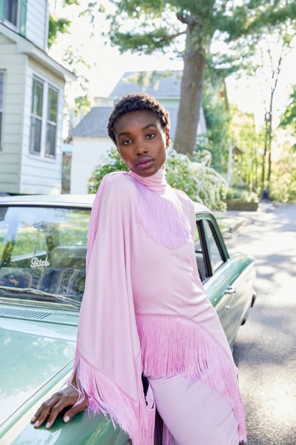 A stylish woman in a pink fringe dress poses against a vintage car in a suburban setting, bathed in soft natural light.