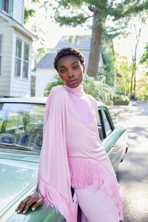 A stylish woman in a pink fringe dress poses against a vintage car in a suburban setting, bathed in soft natural light.