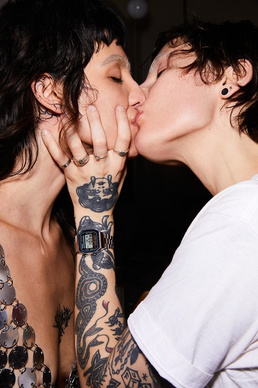 Two individuals with hand tattoos embrace closely, sharing a kiss in a dimly lit room. One is adorned with a silver, sequin-like top.