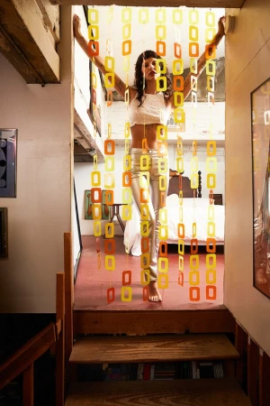 Indoor studio shot of a female model standing in doorway with orange and yellow plastic square links. Bedroom is visible in background.